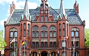 Ein historisches Gebäude aus rotem Backstein mit kunstvollen Verzierungen, Bogenfenstern und mehreren spitzen Türmen. Vor dem Gebäude stehen vier Fahnenmasten mit verschiedenen Flaggen, und zu beiden Seiten rahmen Bäume die Szenerie ein.
