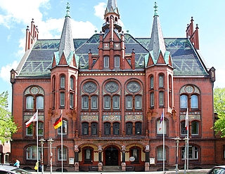 Ein historisches Gebäude aus rotem Backstein mit kunstvollen Verzierungen, Bogenfenstern und mehreren spitzen Türmen. Vor dem Gebäude stehen vier Fahnenmasten mit verschiedenen Flaggen, und zu beiden Seiten rahmen Bäume die Szenerie ein.