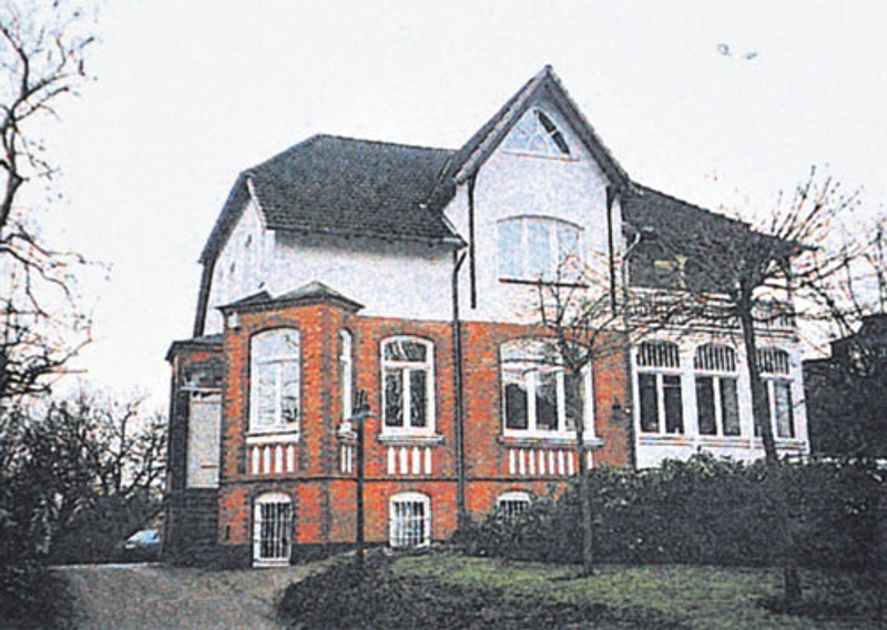 Ein zweistöckiges Haus mit dunklem Dach, weißen oberen Wänden und rotem Backstein im unteren Bereich, mit großen Fenstern und umgeben von Bäumen und Büschen.