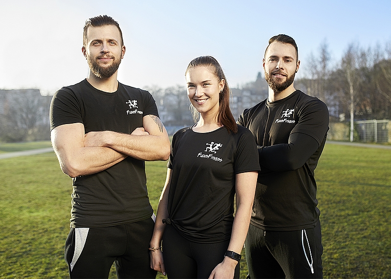 Drei Personen in schwarzen Sportshirts mit dem „Faster Fitness“-Logo stehen draußen auf einer Wiese und lächeln in die Kamera. Die Sonne scheint, und im Hintergrund sind Bäume und Gebäude zu sehen.